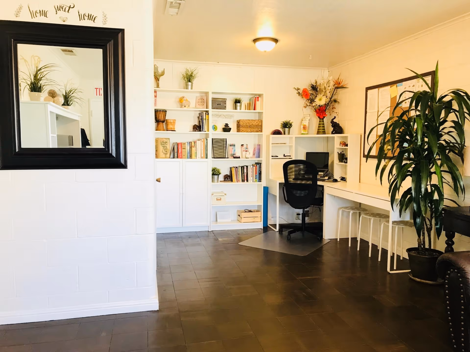 A bright and tidy interior room with white walls and dark flooring. There is a black-framed mirror on the left wall with the words 'home sweet home' above it. In the background, there are white shelves filled with books, plants, and decorative items. A desk with a computer and a black office chair is positioned against the right wall, accompanied by three white stools. A large potted plant is also visible on the right side of the room.