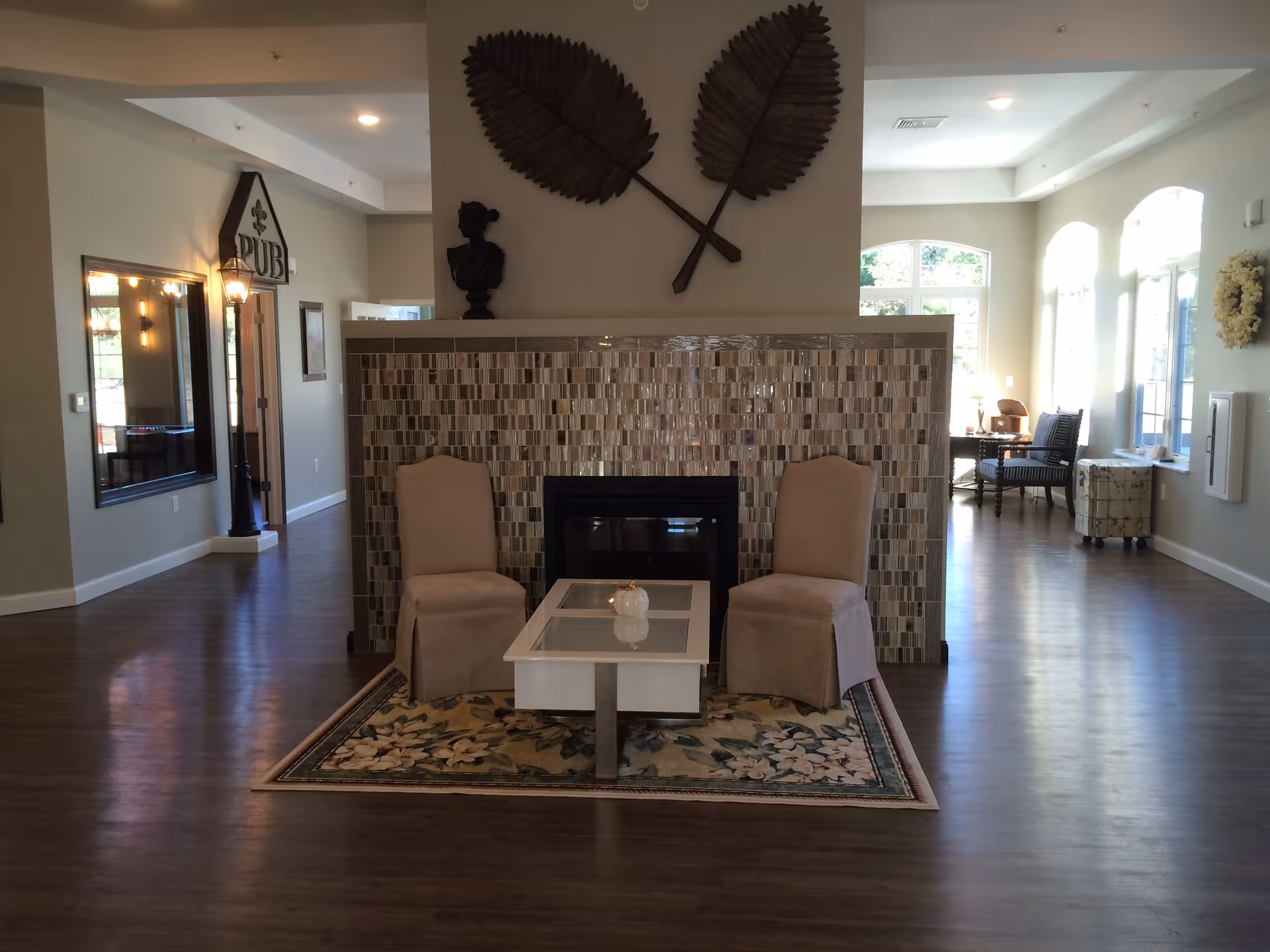 A cozy interior common area with two beige upholstered chairs and a white coffee table placed on a floral rug in front of a tiled fireplace. Above the fireplace are two large decorative leaves mounted on the wall. The room has wooden flooring, large windows letting in natural light, and additional seating areas visible in the background.
