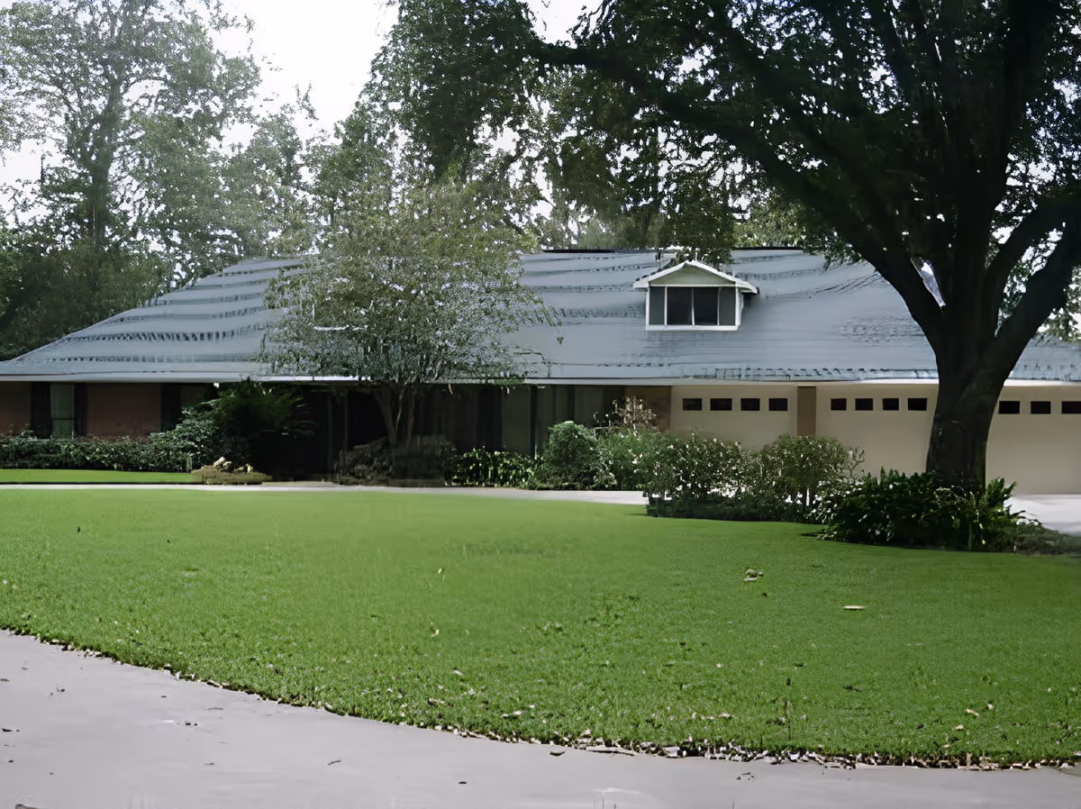 Single-story house with a wide roof, three-car garage, large green lawn and mature trees in front.