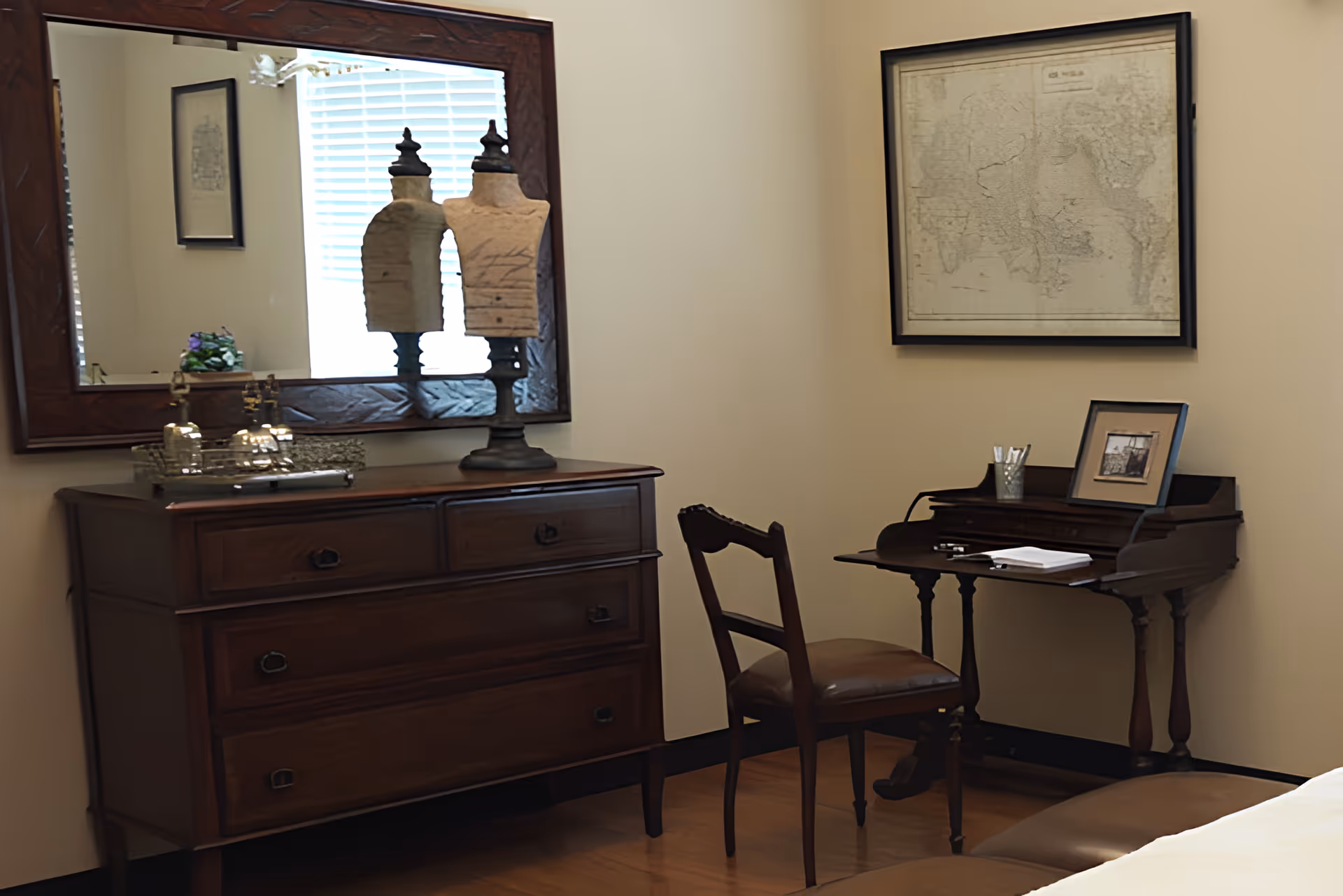 A bedroom corner with a wooden dresser topped by a large mirror and decorative items, and a small writing desk with a chair and framed art on the wall.