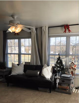 Sunlit living room with a black sofa and white pillows, side tables, a small Christmas tree, and large multi-pane windows.