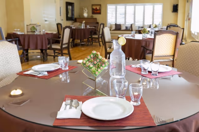 A bright dining room with round tables set with plates, glasses, a water carafe, and floral centerpieces.