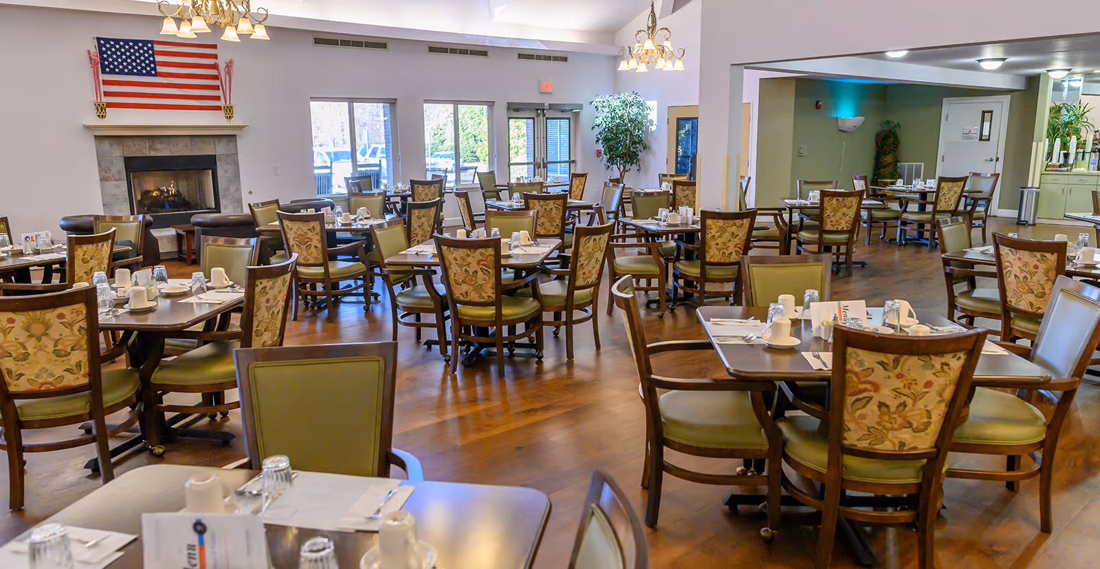 Large senior living dining room with many set tables and chairs, a fireplace with an American flag above it, and glass doors to an outdoor area.