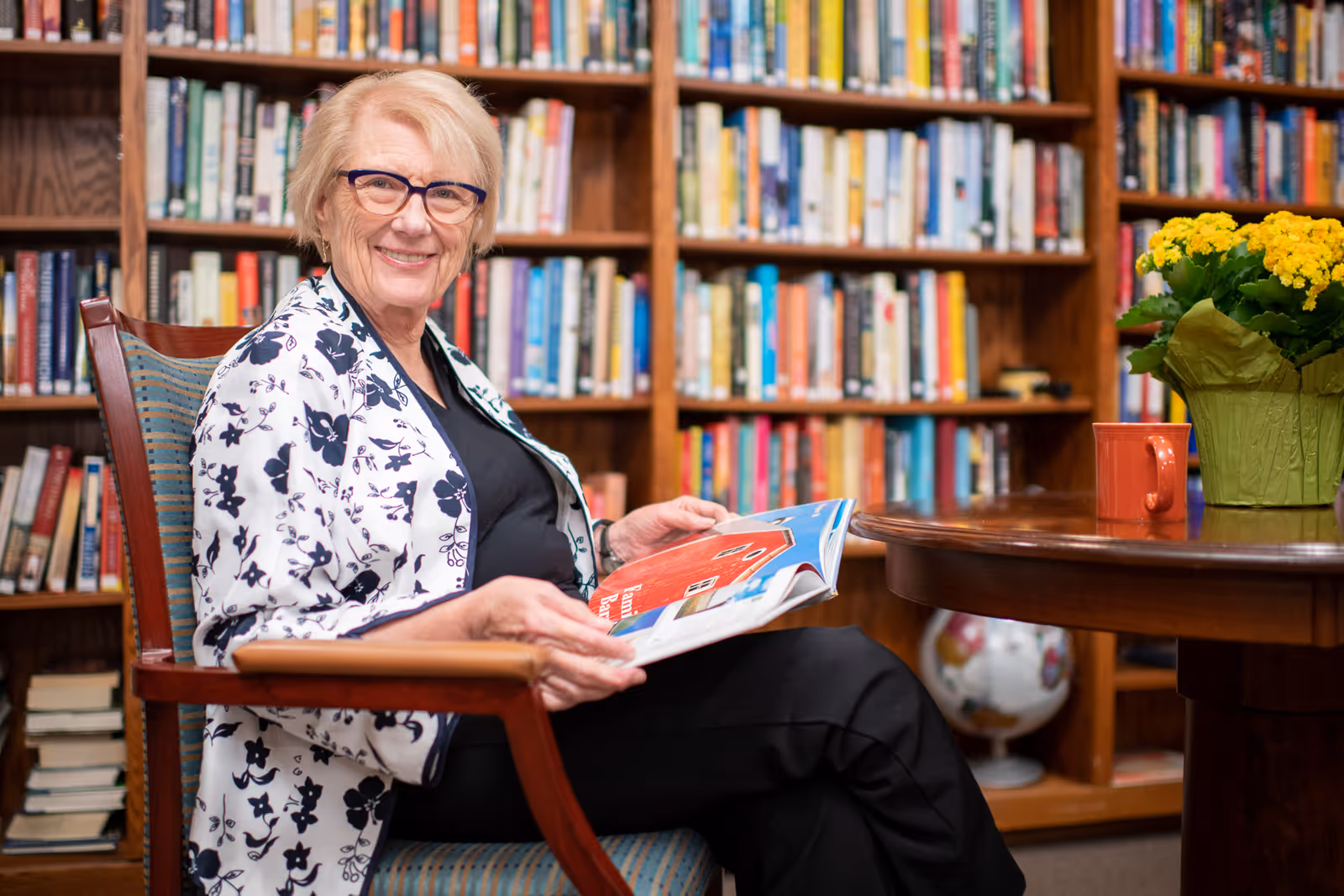 An elderly woman with short blonde hair and glasses is sitting in a wooden chair with a patterned cushion, holding an open magazine. She is smiling and wearing a white jacket with a black floral pattern over a black top. Behind her is a large wooden bookshelf filled with books. A round wooden table with a red mug and a green pot with yellow flowers is next to her.