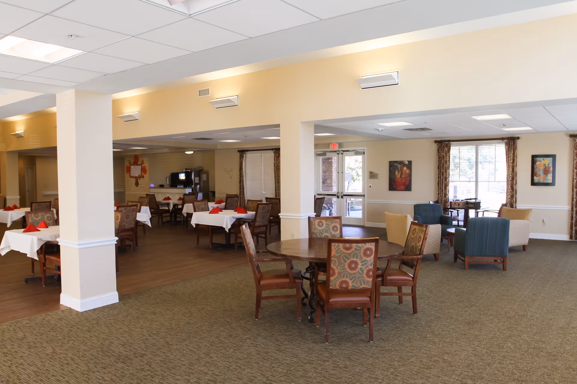 A spacious dining and lounge area in an assisted living facility. The room features several tables with chairs, some tables set with white tablecloths and red napkins. There are comfortable armchairs arranged near large windows with patterned curtains, and framed artwork on the walls. The space is well-lit with ceiling lights and wall sconces, and has a mix of carpeted and wood flooring.