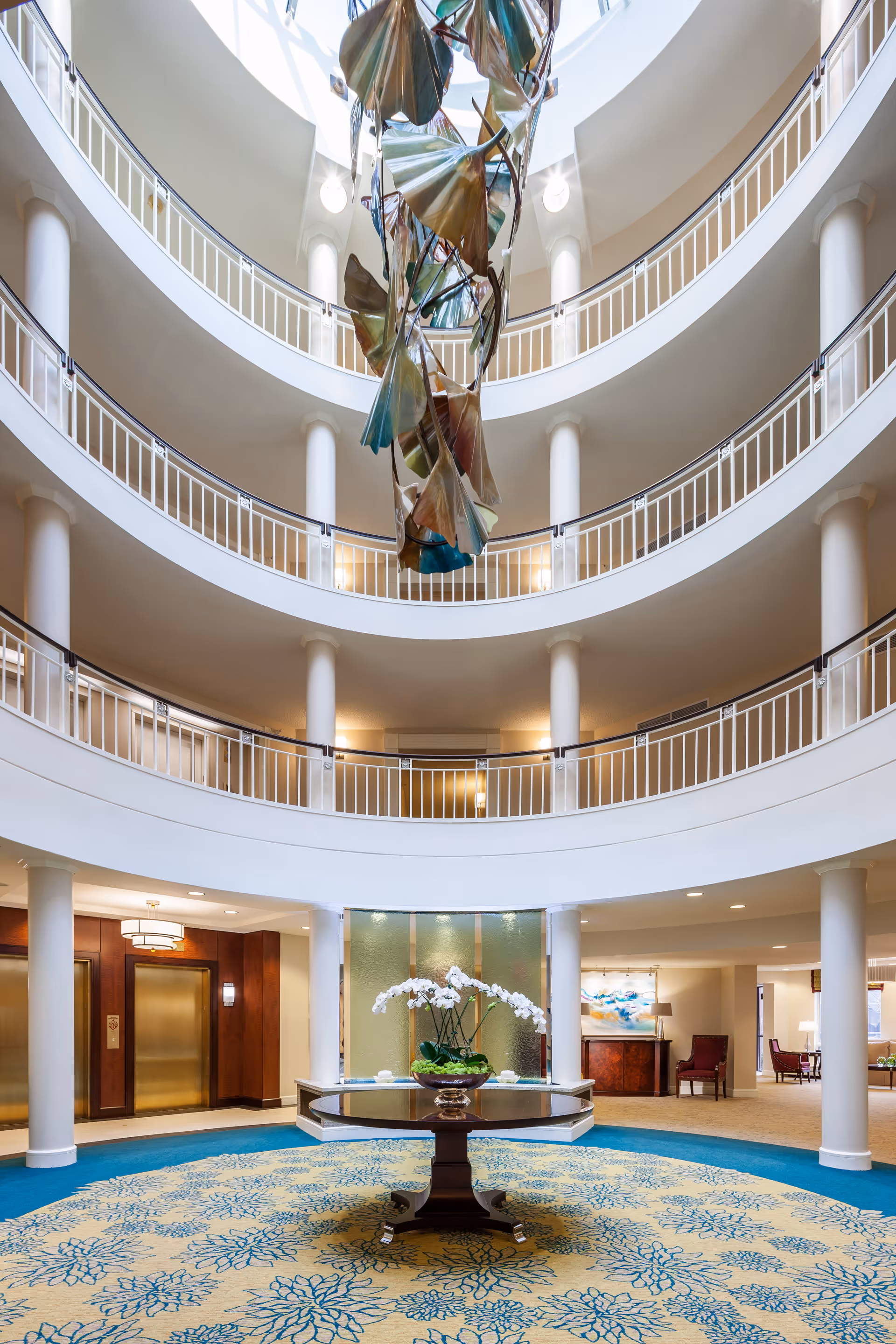 Multi-story circular atrium with a central table holding white orchids, a hanging sculpture from a skylight, and surrounding balcony levels.