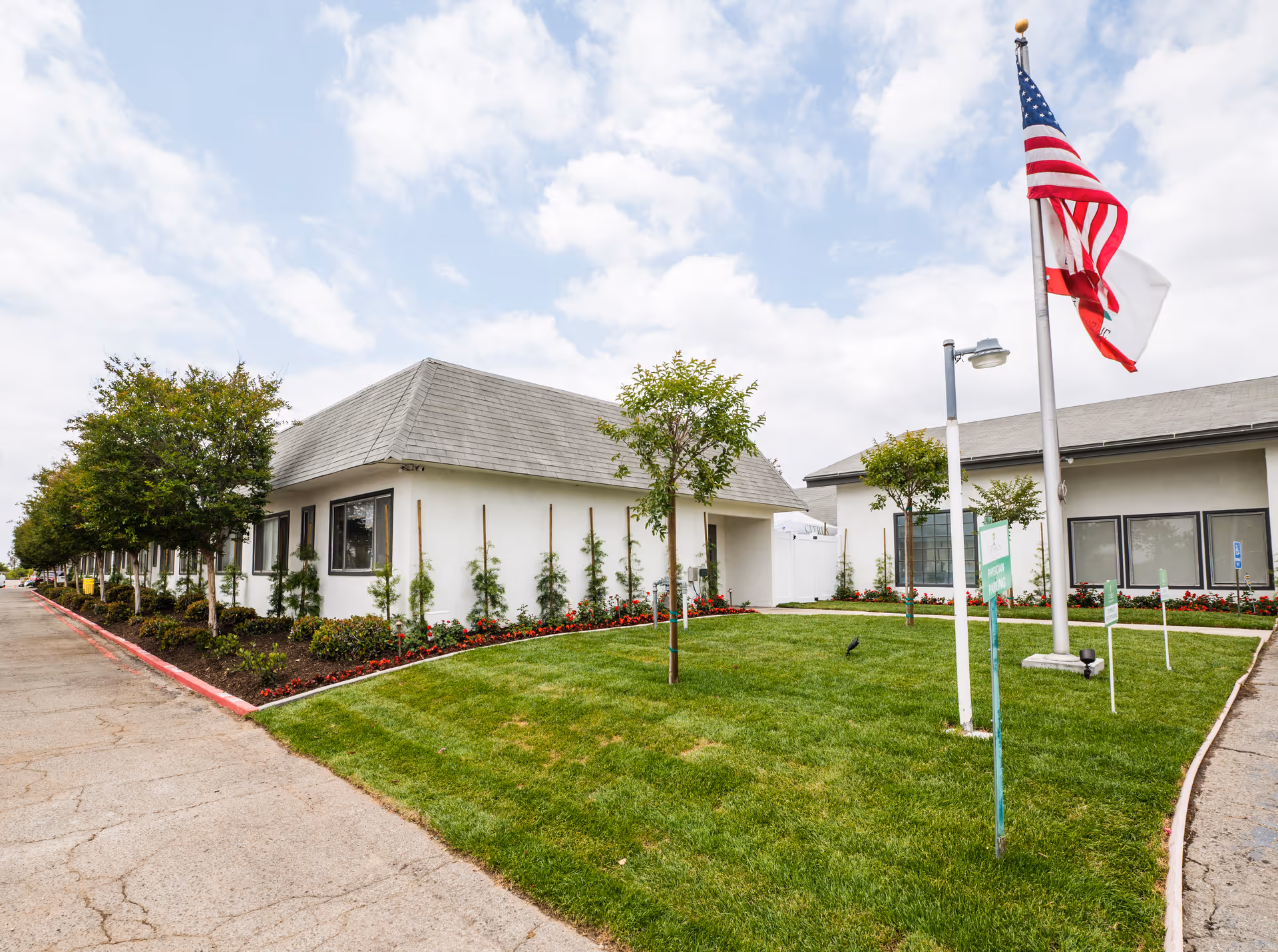 Exterior view of a single-story nursing center building with a gray roof, white walls, and several windows. There is a well-maintained lawn with small trees and flower beds in front of the building. Two flagpoles with an American flag and another flag are visible on the lawn. The sky is partly cloudy.