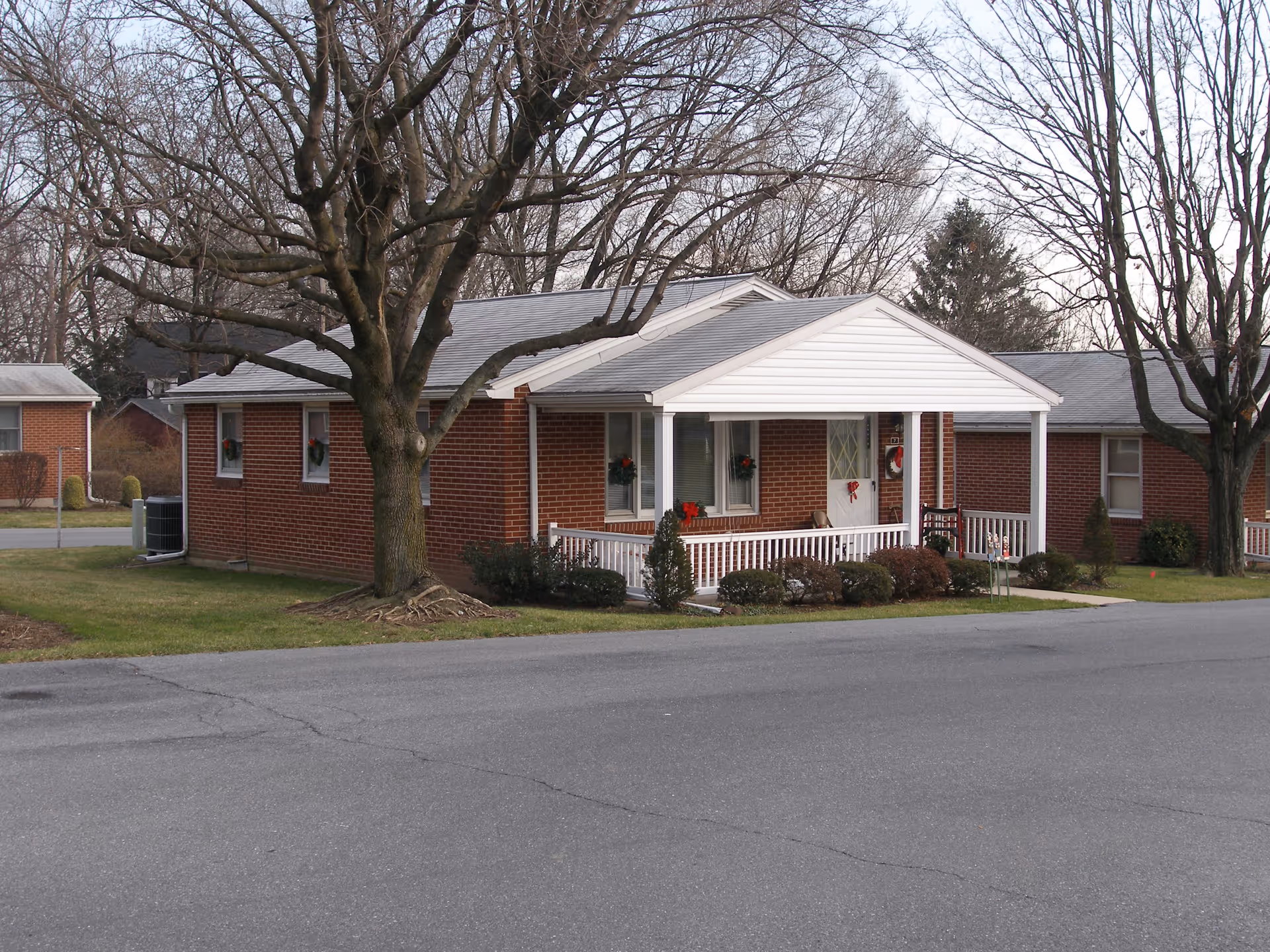 A single-story brick building with a white porch roof and railing, decorated with holiday wreaths on the windows and door. There are leafless trees and shrubs around the building, and a paved road in the foreground.