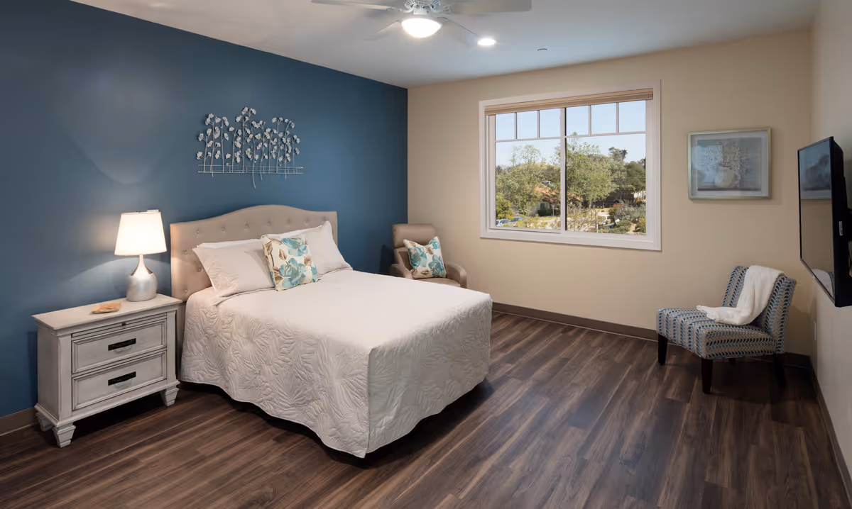 A neatly arranged bedroom with a bed covered in a white quilt and floral pillows, a nightstand with a lamp, a beige armchair with a floral pillow, a patterned chair with a white throw, a large window showing trees outside, and a wall-mounted TV.