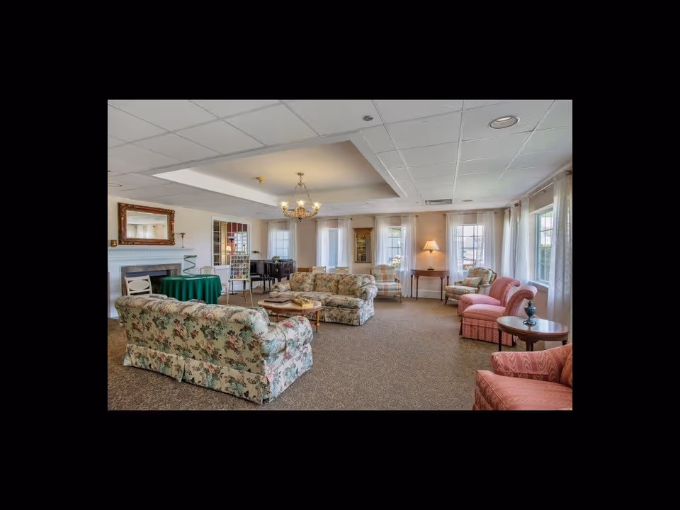 A spacious living room with floral patterned sofas and armchairs arranged around a coffee table. The room has multiple windows with sheer white curtains allowing natural light to fill the space. There is a chandelier hanging from the ceiling, a fireplace with a large mirror above it, a green-covered round table, and a piano in the background. The carpet is brown, and the walls are light-colored, creating a warm and inviting atmosphere.