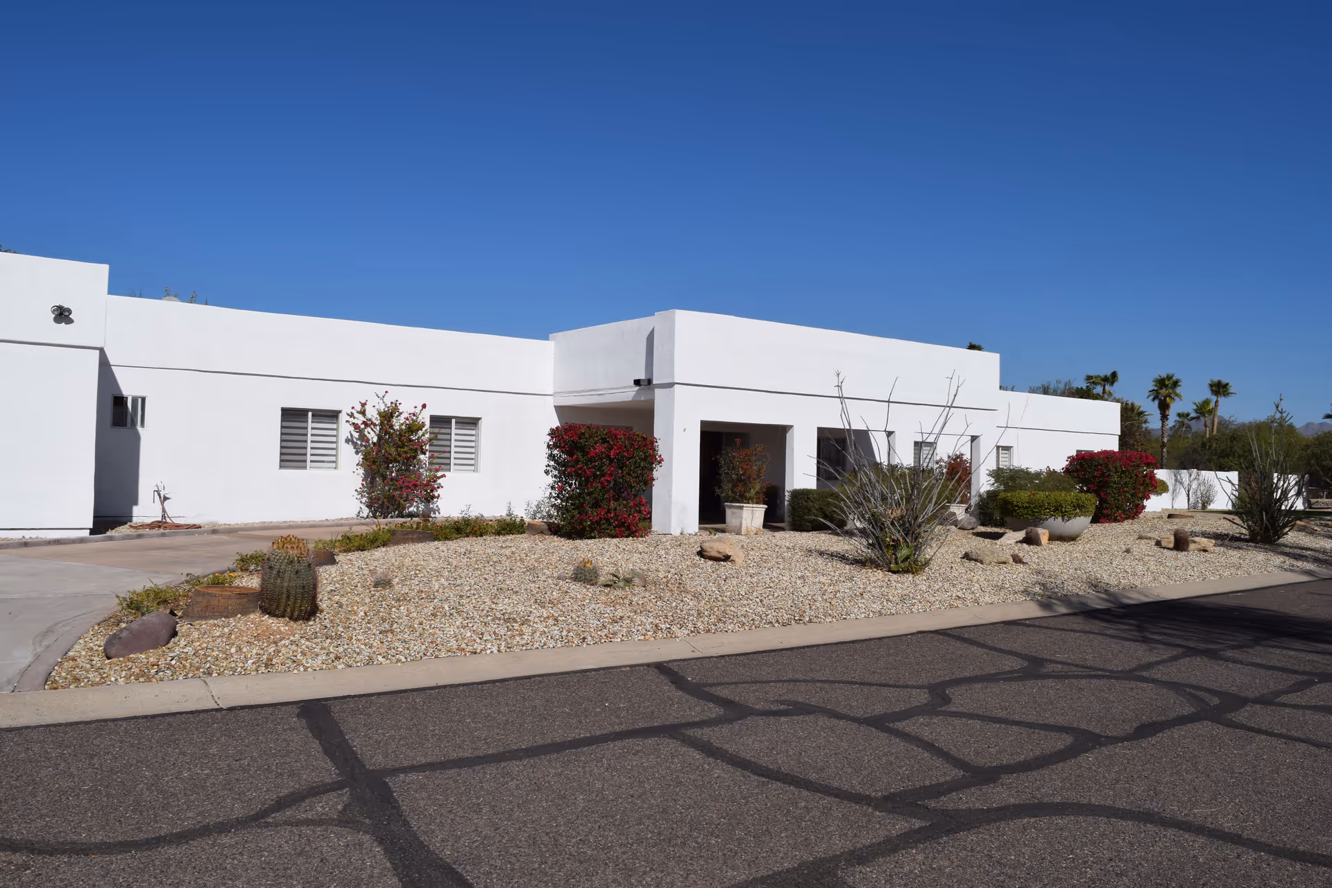 Exterior view of a single-story white building with a flat roof, desert landscaping with rocks, cacti, and bushes, and a clear blue sky overhead.