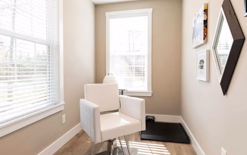 A small, bright room with beige walls and wood flooring featuring a white cushioned chair on a metal base, two windows with white blinds letting in natural light, a black mat on the floor, and three framed pieces of wall art including a diamond-shaped mirror.