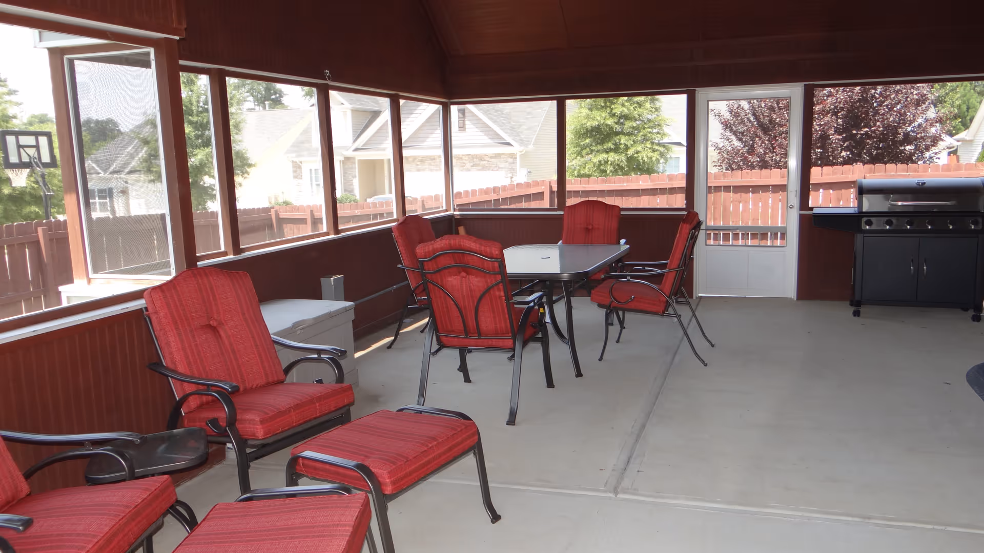 A screened-in porch area with red cushioned chairs and a matching ottoman, a glass-top table with four red cushioned chairs around it, and a black grill against the far wall. Outside the porch, there is a wooden fence and neighboring houses visible.