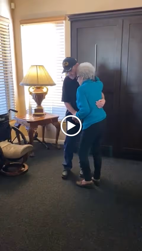An elderly man and woman standing and embracing in a room with a wooden cabinet, a side table with a lamp, a wheelchair, and window blinds.