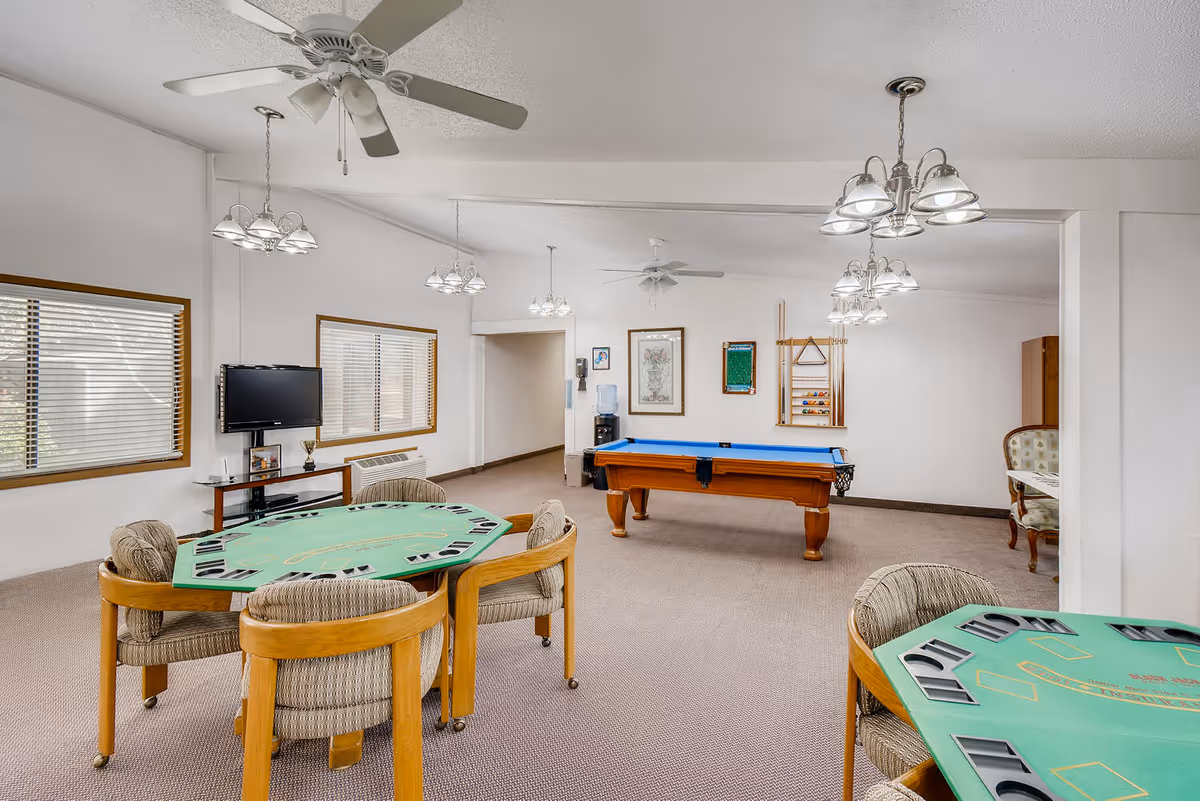 A spacious senior living community game room with two card tables surrounded by chairs, a pool table with a blue felt top, a wall-mounted TV, and framed artwork on the walls. The room has beige carpeting, white walls, ceiling fans, and multiple hanging light fixtures.