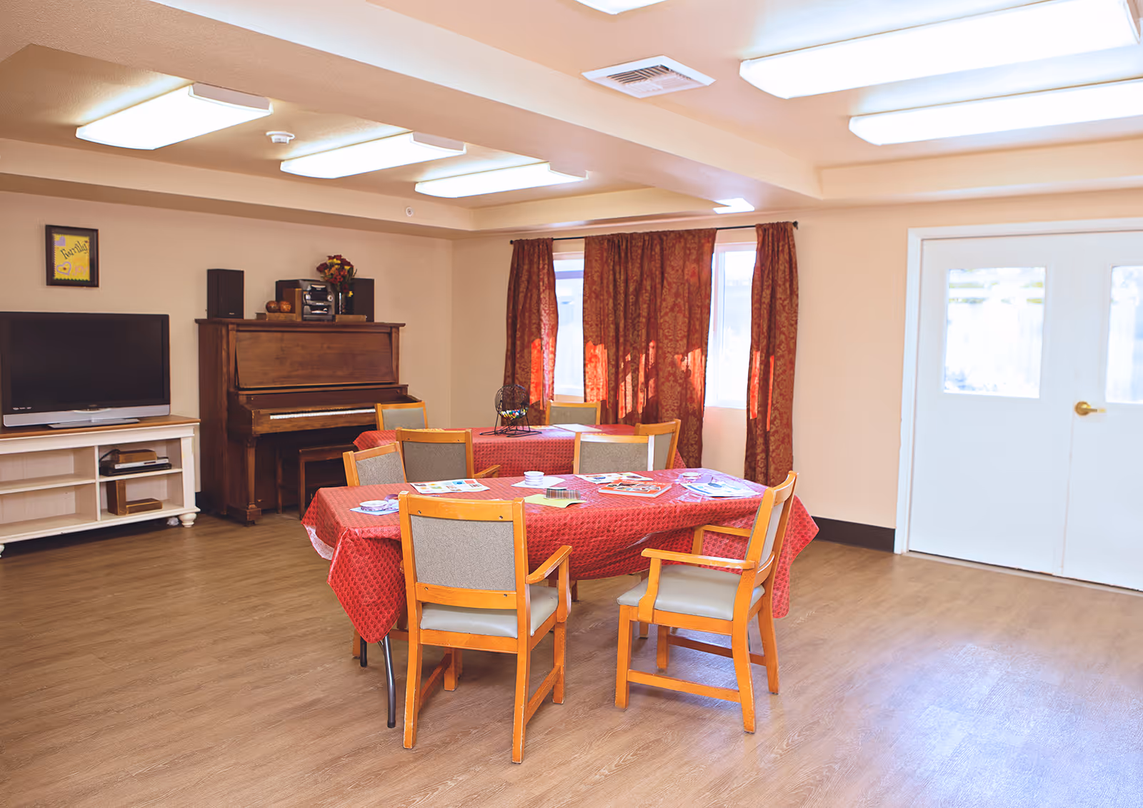 A bright communal dining/activity room with a red tablecloth-covered table and wooden chairs, a piano and TV against the wall, and double doors.