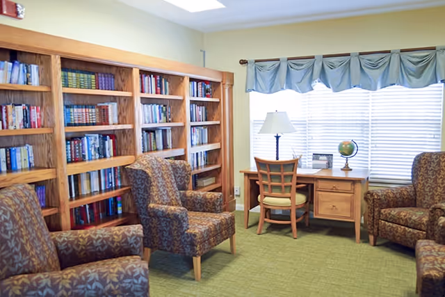 A cozy reading room with wooden bookshelves, patterned upholstered armchairs, and a desk with a lamp and globe by a window.