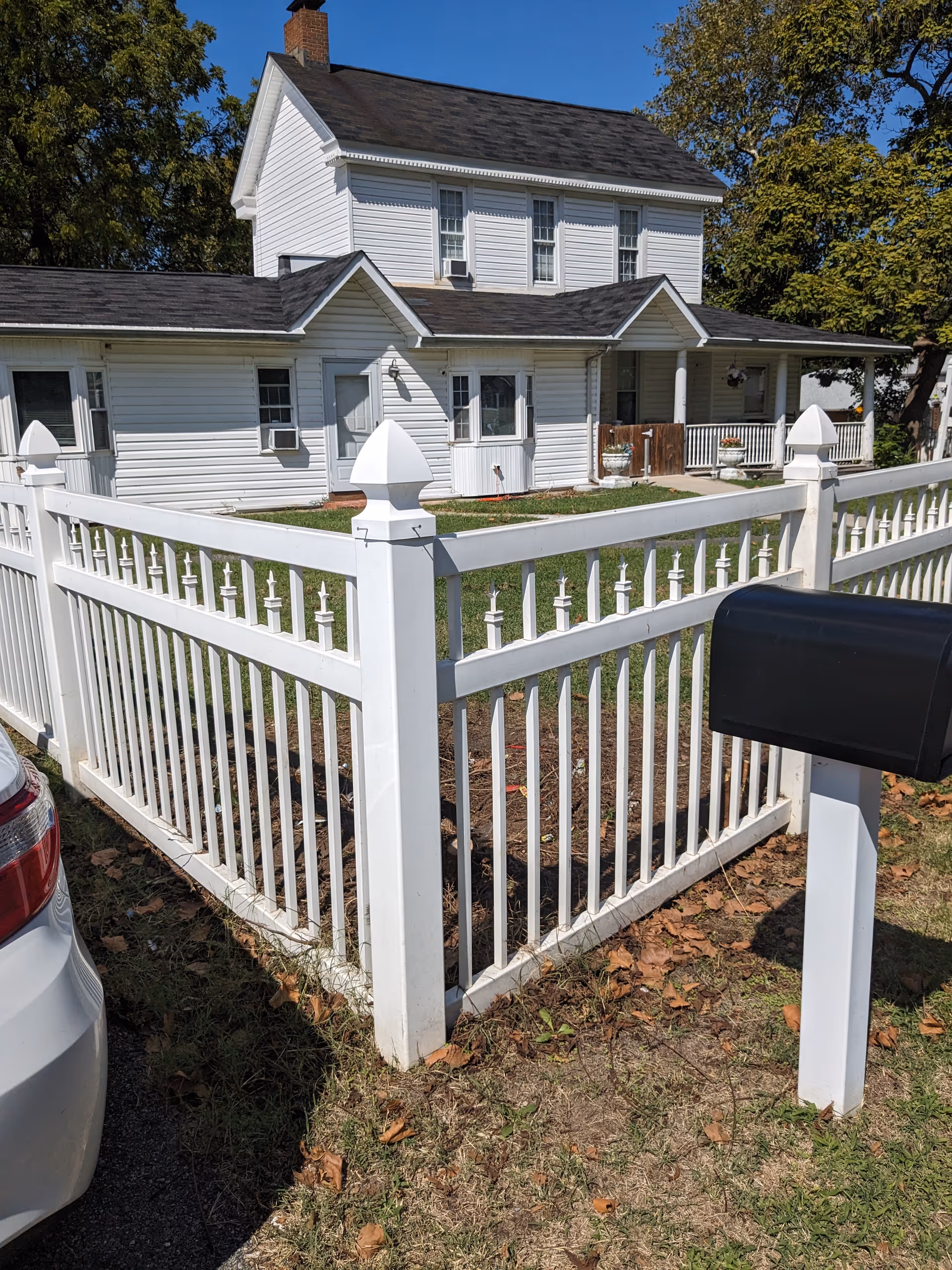 White two-story house with a dark shingled roof, surrounded by a white picket fence. There is a black mailbox on a white post near the fence and a white car partially visible on the left side. The yard has green grass and some fallen leaves, with trees in the background.
