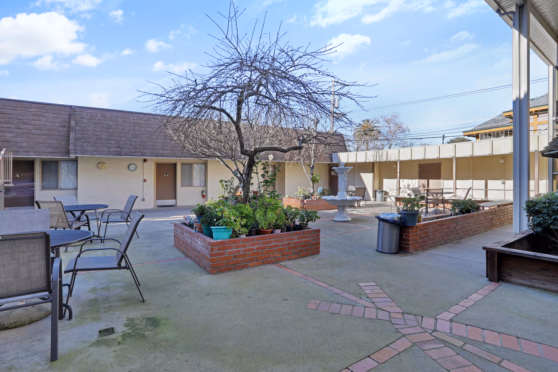 Sunlit courtyard with a central tree and fountain, brick planters and outdoor seating at an assisted living facility.