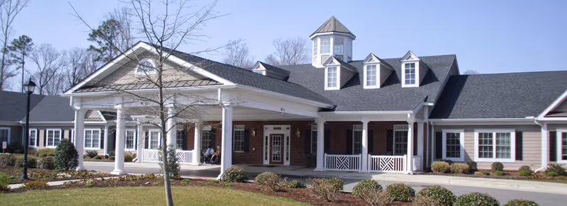 Front exterior view of Spring Arbor of Williamsburg, a single-story building with a covered entrance supported by white columns, brick and siding facade, multiple windows, and a cupola on the roof. The surrounding area includes landscaped bushes and a small tree.
