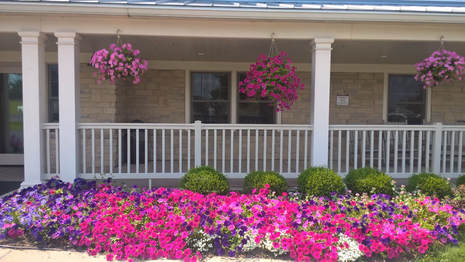 Front porch area of a building with white columns and railing, stone walls, and three hanging baskets filled with pink flowers. Below the porch, there is a garden bed with vibrant pink, purple, and white flowers along with neatly trimmed green bushes.