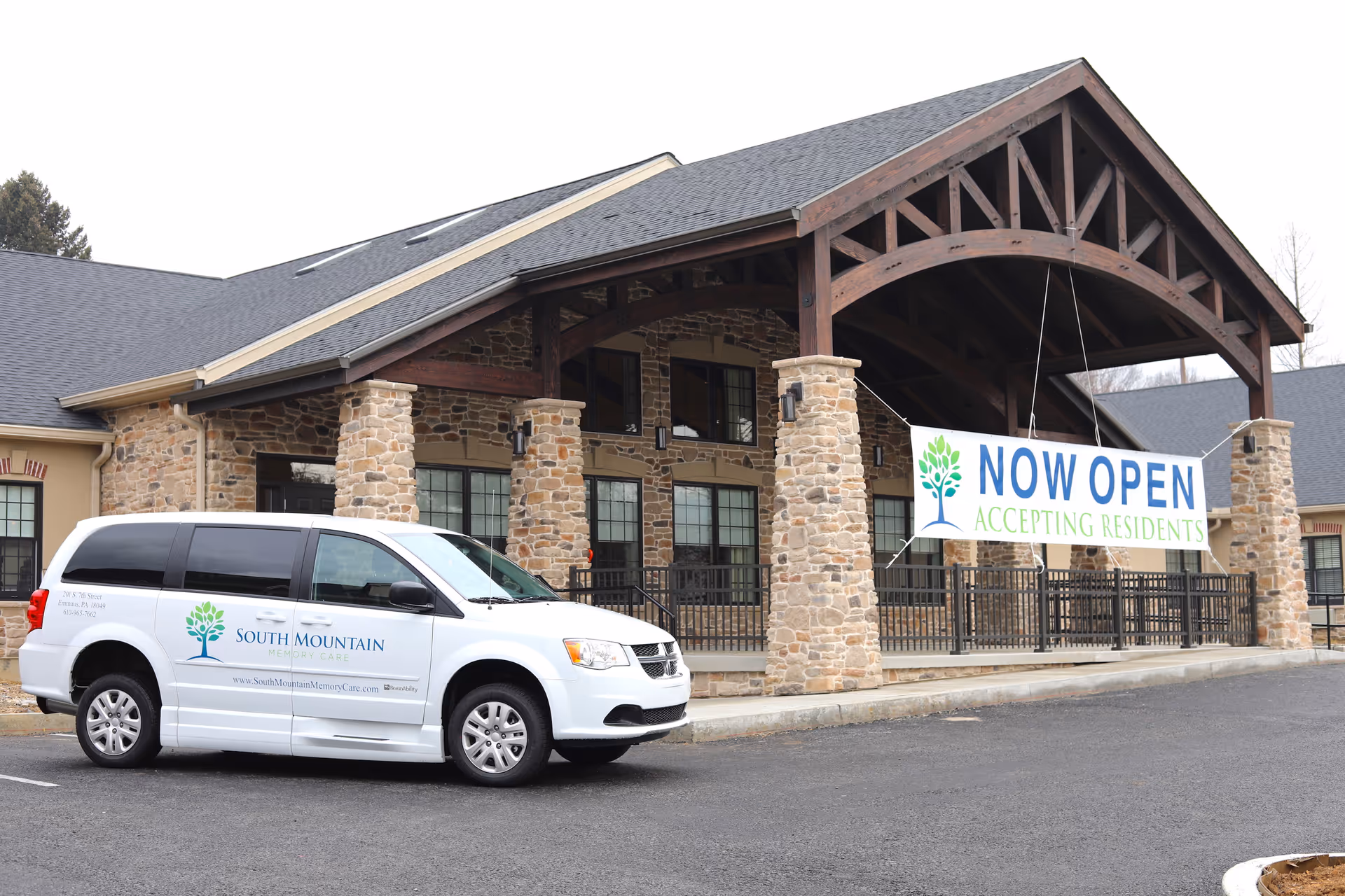 Front entrance of South Mountain Memory Care with a covered stone porte-cochere, a 'NOW OPEN Accepting Residents' banner, and a branded white van parked in front.