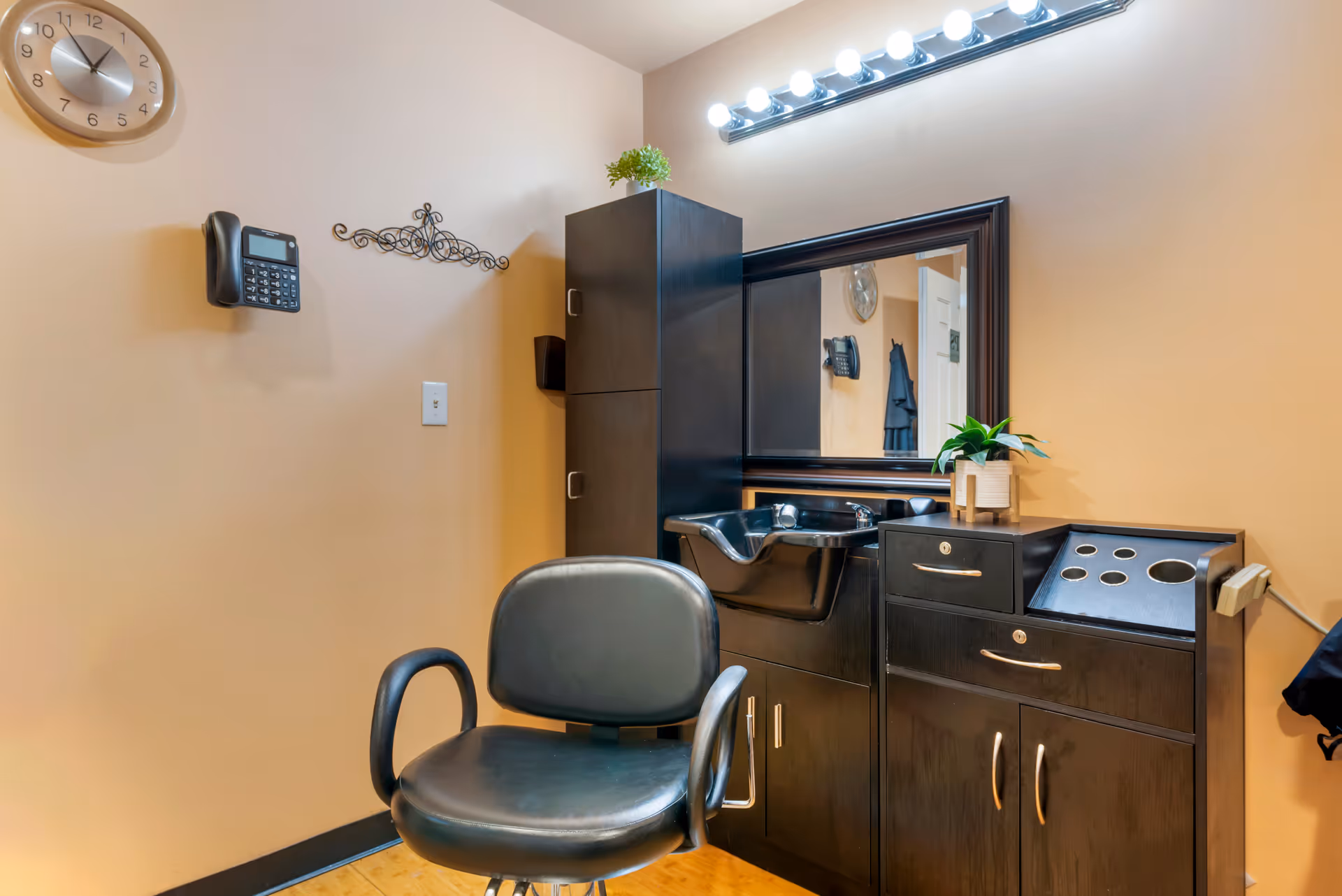 A salon styling station with a black salon chair, wash sink, mirror, cabinetry and vanity lights in an interior room.