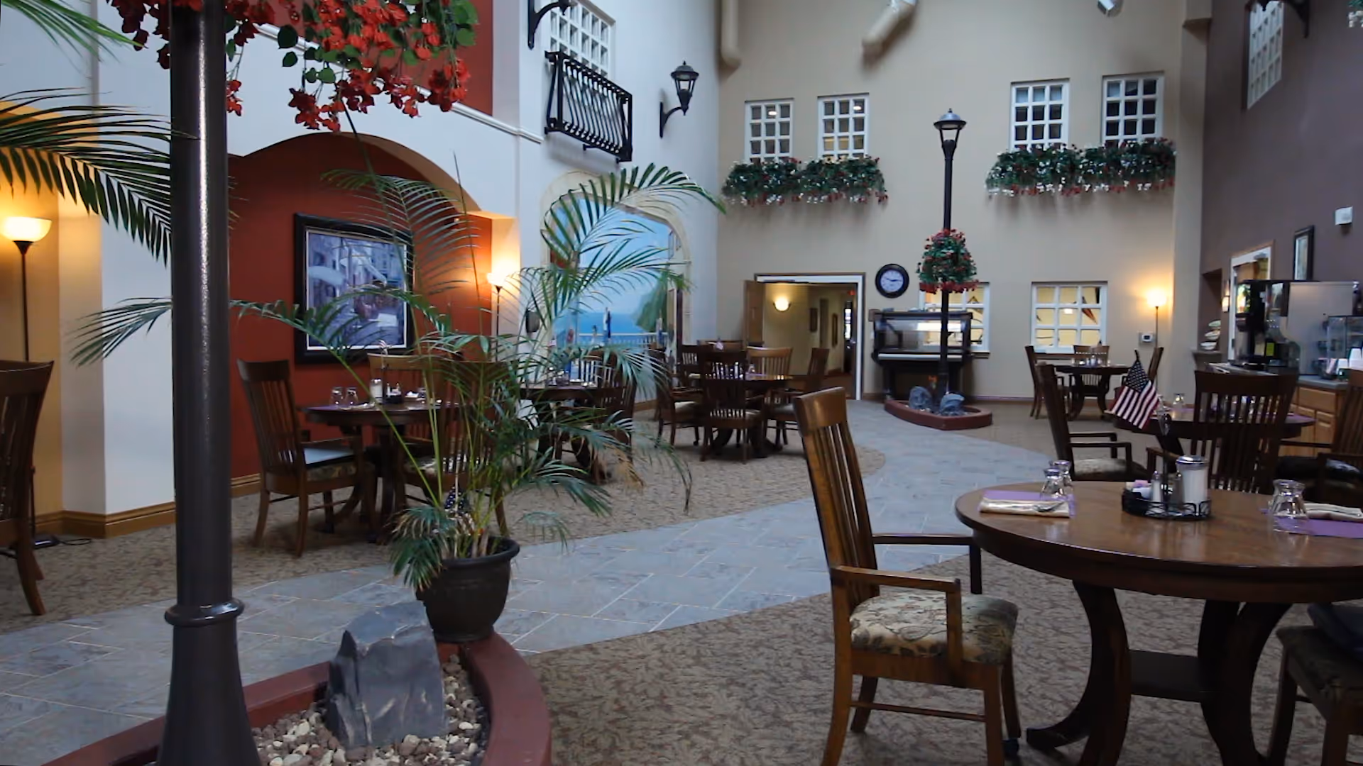 Interior view of a dining area in Sherwood Lodge Assisted Living with round wooden tables and chairs, decorative plants, wall paintings, and a high ceiling with windows. The space is warmly lit with lamps and decorated with hanging flower baskets and a small American flag on one table.