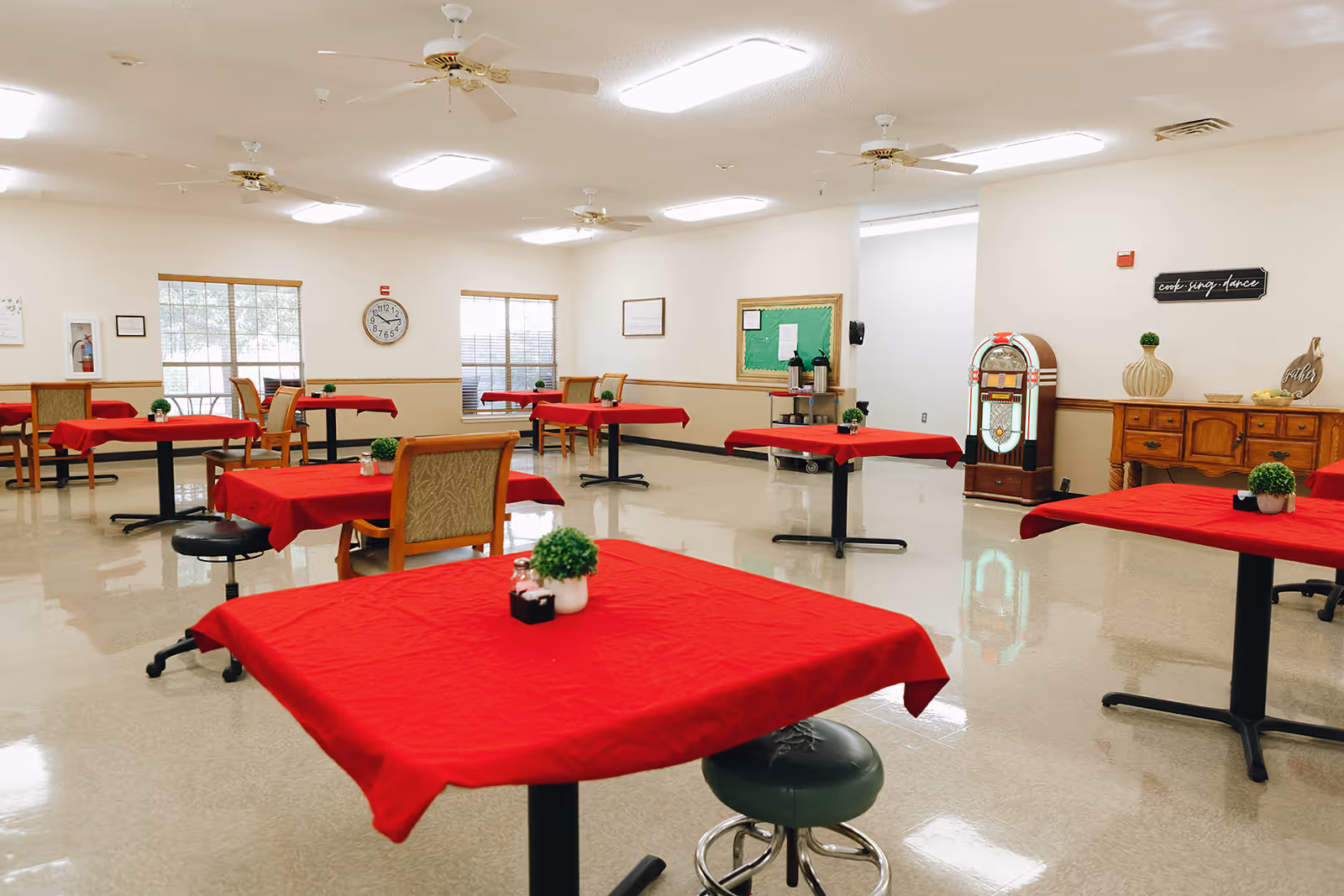 A spacious dining room with multiple tables covered in red tablecloths, each adorned with small potted plants. The room has beige walls, large windows letting in natural light, ceiling fans, and a vintage jukebox near a wooden sideboard. The floor is shiny and reflective, and there are chairs and stools around the tables.