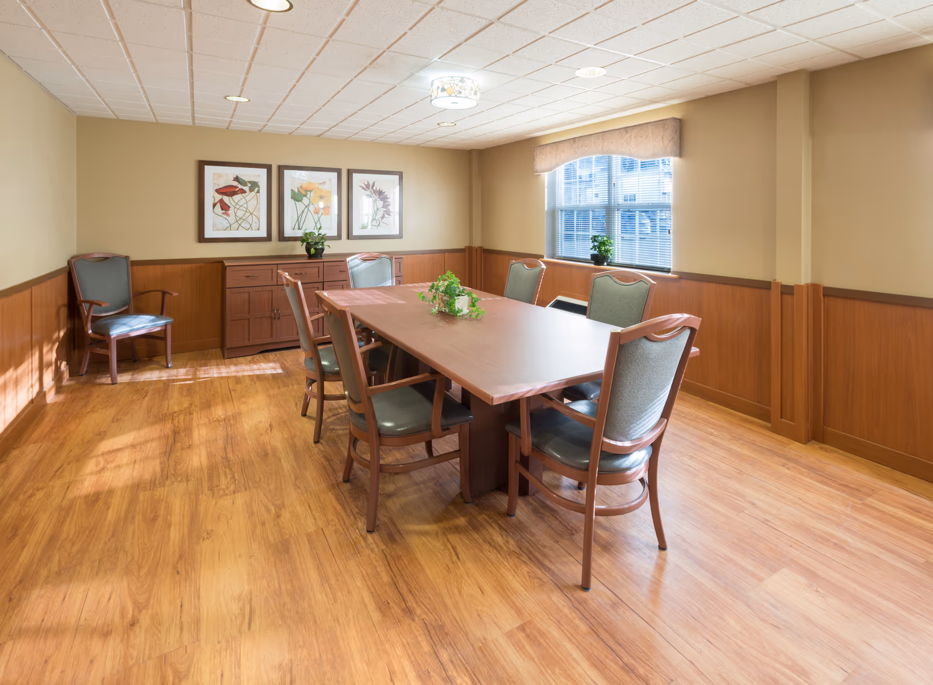 A well-lit dining room with a rectangular wooden table surrounded by six cushioned chairs. The room has wooden flooring and wood paneling on the lower half of the walls. There are three framed floral artworks on the wall above a wooden sideboard, and a window with blinds and a valance letting in natural light. A small green plant is placed on the table and another on the sideboard.