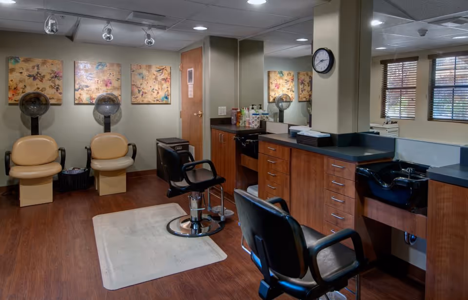 Interior of a salon area in a senior living facility with two beige hair dryer chairs against the wall, two black salon chairs in front of mirrors and countertops with hair care products, and a black hair washing sink. The room has wooden flooring, decorative wall art, and windows with blinds letting in natural light.