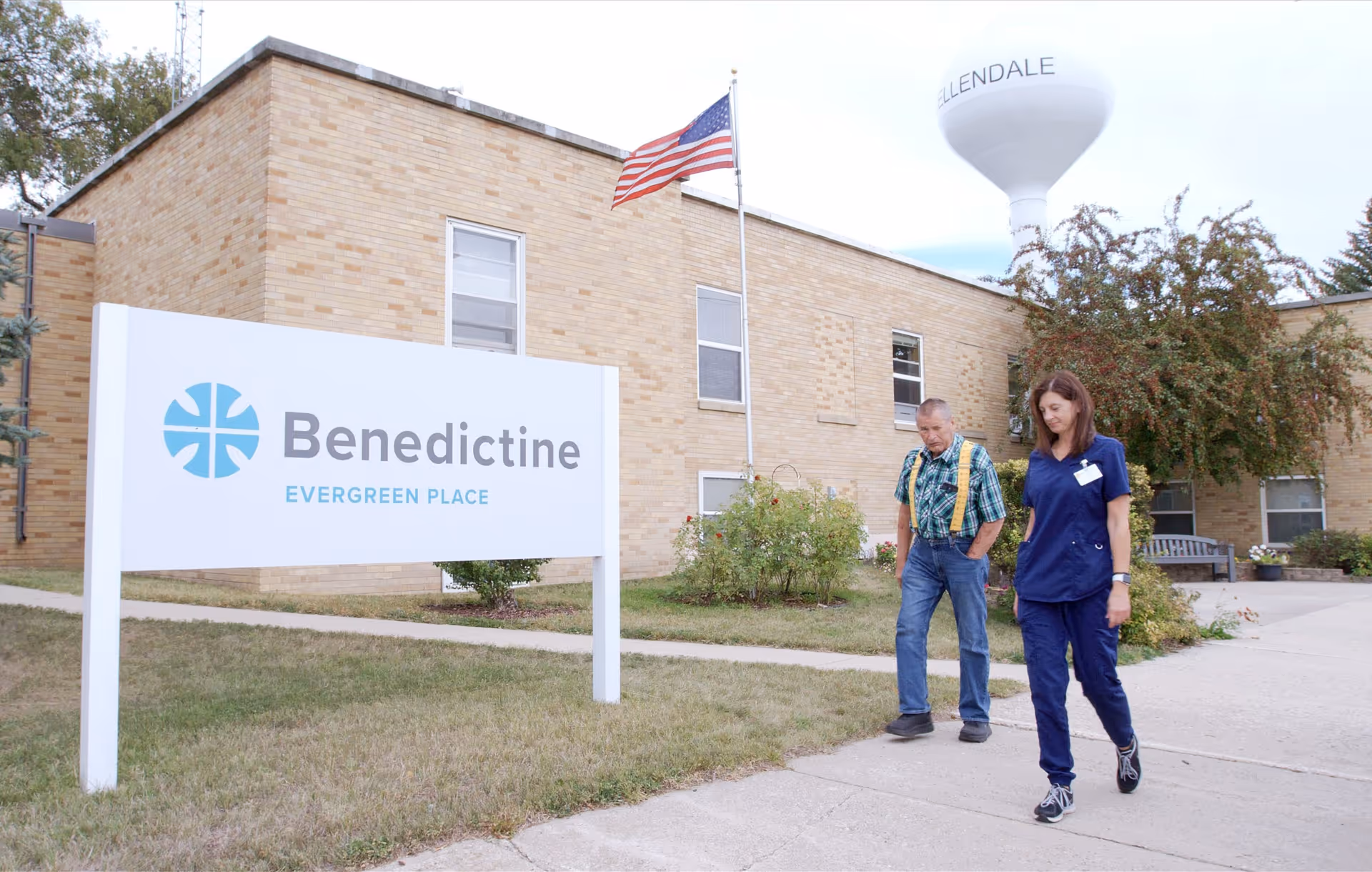 Two people walk past a white sign reading 'Benedictine Evergreen Place' in front of a low brick building with an American flag and a water tower in the background.