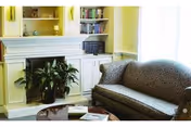 A cozy living room area with a patterned sofa, a coffee table with books and papers, a large green plant, built-in white shelves filled with books, and a white fireplace mantel. The walls are painted yellow and there is natural light coming through a window with sheer white curtains.