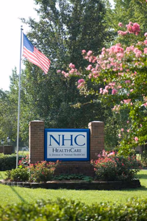 Exterior view of NHC Healthcare Sumter sign with American flag and pink blooming shrubs.