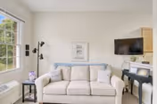 A bright living room with a beige sofa adorned with two white pillows, a small side table with a purple flower vase, a wall-mounted flat-screen TV, a black piano, and a large window letting in natural light.