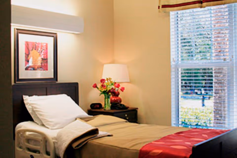 A neatly made single bed with beige and red bedding in a room with beige walls. There is a wooden headboard, a nightstand with a lamp, a vase of flowers, and an alarm clock. A window with white blinds lets in natural light and shows greenery outside. A framed picture hangs on the wall above the bed.