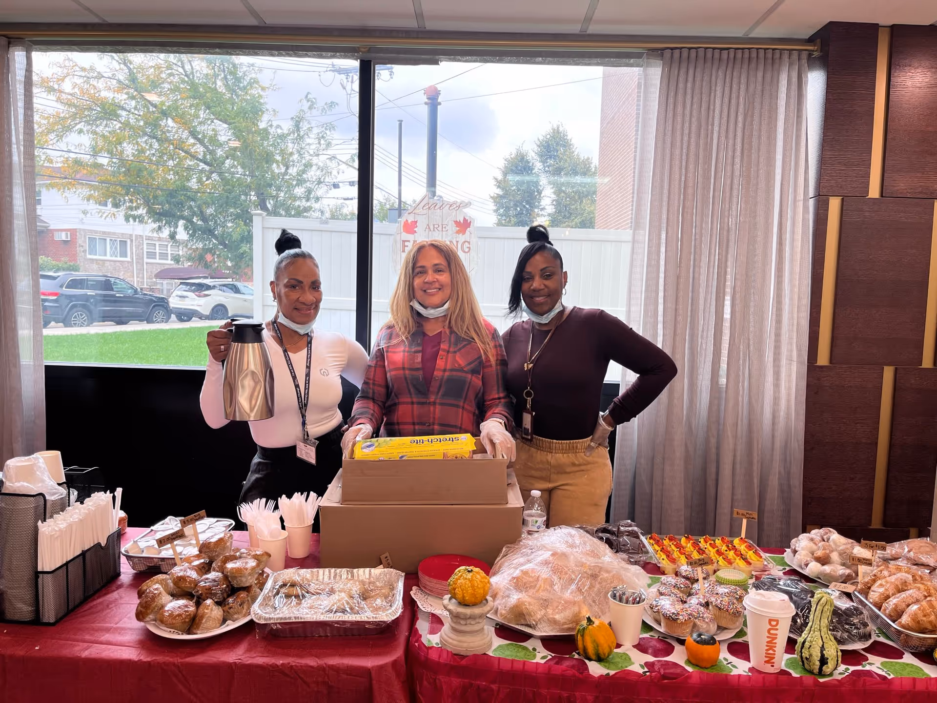 Three women standing behind a buffet table filled with pastries, coffee, and snacks in a dining area.