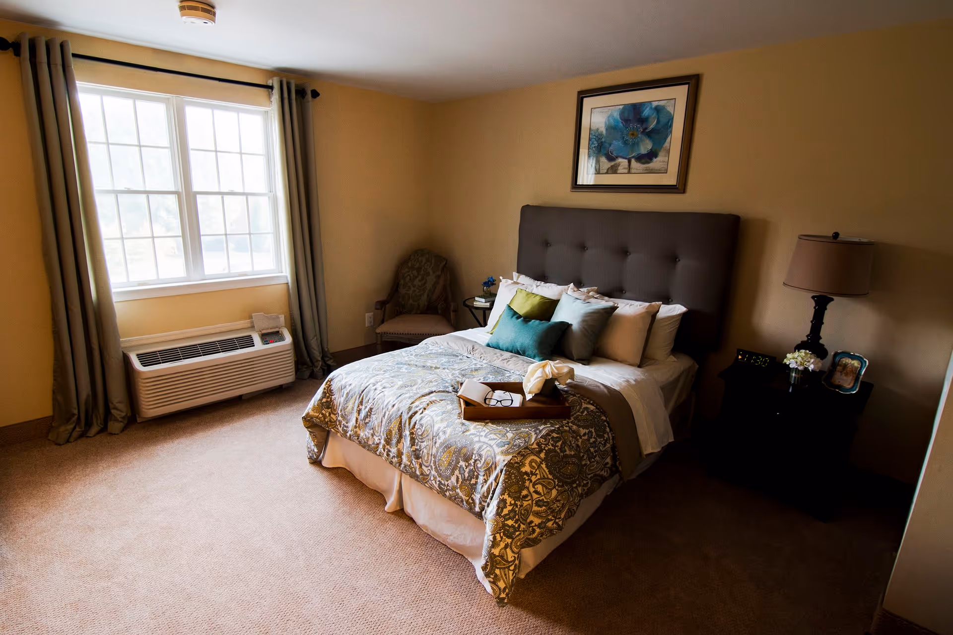A cozy bedroom with a large window covered by beige curtains, a bed with a tufted dark gray headboard, multiple pillows, and a patterned bedspread. There is a wooden nightstand with a lamp, a digital clock, a small flower vase, and a framed photo. A cushioned chair is placed in the corner near the window.