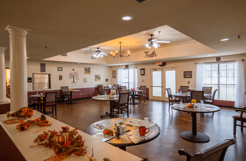 A spacious dining room in an assisted living facility with several round tables set with placemats, utensils, cups, and small floral centerpieces. The room has wooden floors, large windows with white curtains, ceiling fans, and warm lighting. Autumn-themed decorations are placed on a counter in the foreground. The word 'Faith' is displayed on the wall above the exit door.