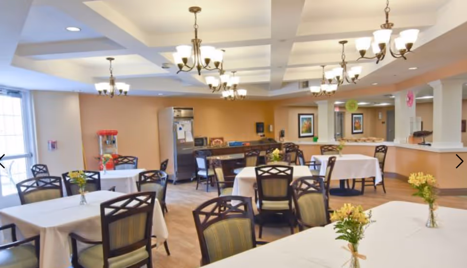 A bright dining room with multiple tables covered in white tablecloths, each adorned with small flower arrangements. The room features several chairs with patterned backs, a popcorn machine near the back wall, and a counter area with columns. The ceiling has recessed lighting and hanging chandeliers, and the walls are painted in warm tones with framed artwork.