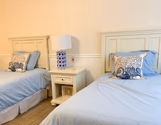 A bright bedroom with two twin beds featuring light blue bedding and decorative pillows with a seashell design. Between the beds is a white nightstand with a blue and white patterned lamp. The walls are light-colored with a white chair rail molding.
