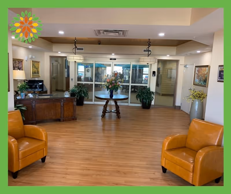 A spacious and well-lit reception area with wooden flooring, two orange leather armchairs, a wooden reception desk with a computer, and a round table with a floral arrangement in the center. The background features glass sliding doors and potted plants on either side. The walls are decorated with framed paintings and a large vase with yellow flowers.