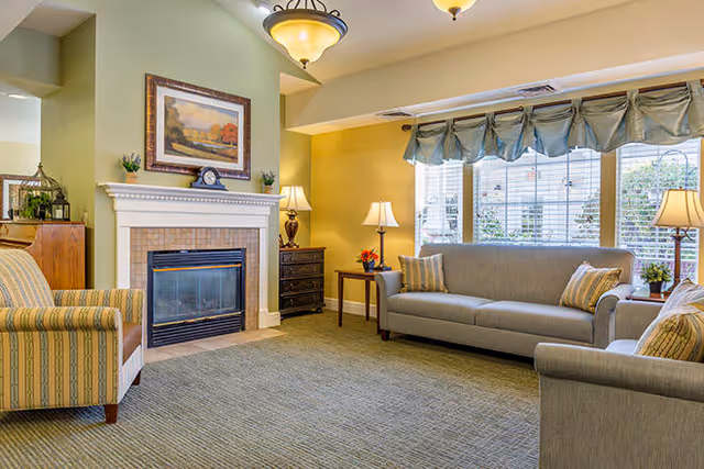 A cozy living room with a fireplace, two gray sofas with striped pillows, a striped armchair, two side tables with lamps, a framed painting above the fireplace, and large windows with curtains letting in natural light.