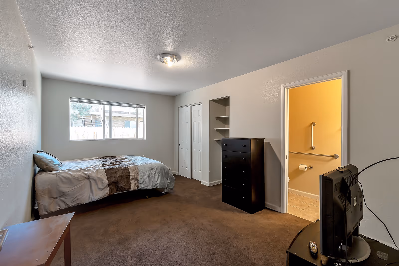 A simple bedroom with a single bed against the left wall, a window above the bed letting in natural light, a black dresser and open shelving on the right wall, and an open door leading to a bathroom with grab bars. A TV is placed on a stand in the foreground on the right side.