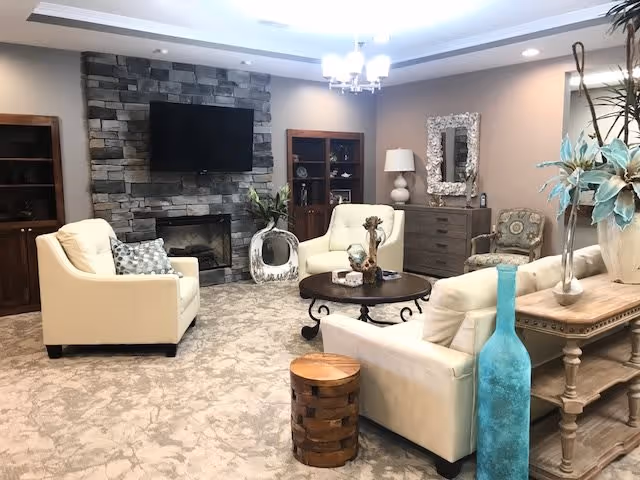 Cozy retirement community lounge with cream sofas arranged around a round coffee table, a stone fireplace with a mounted TV, and decorative furnishings.