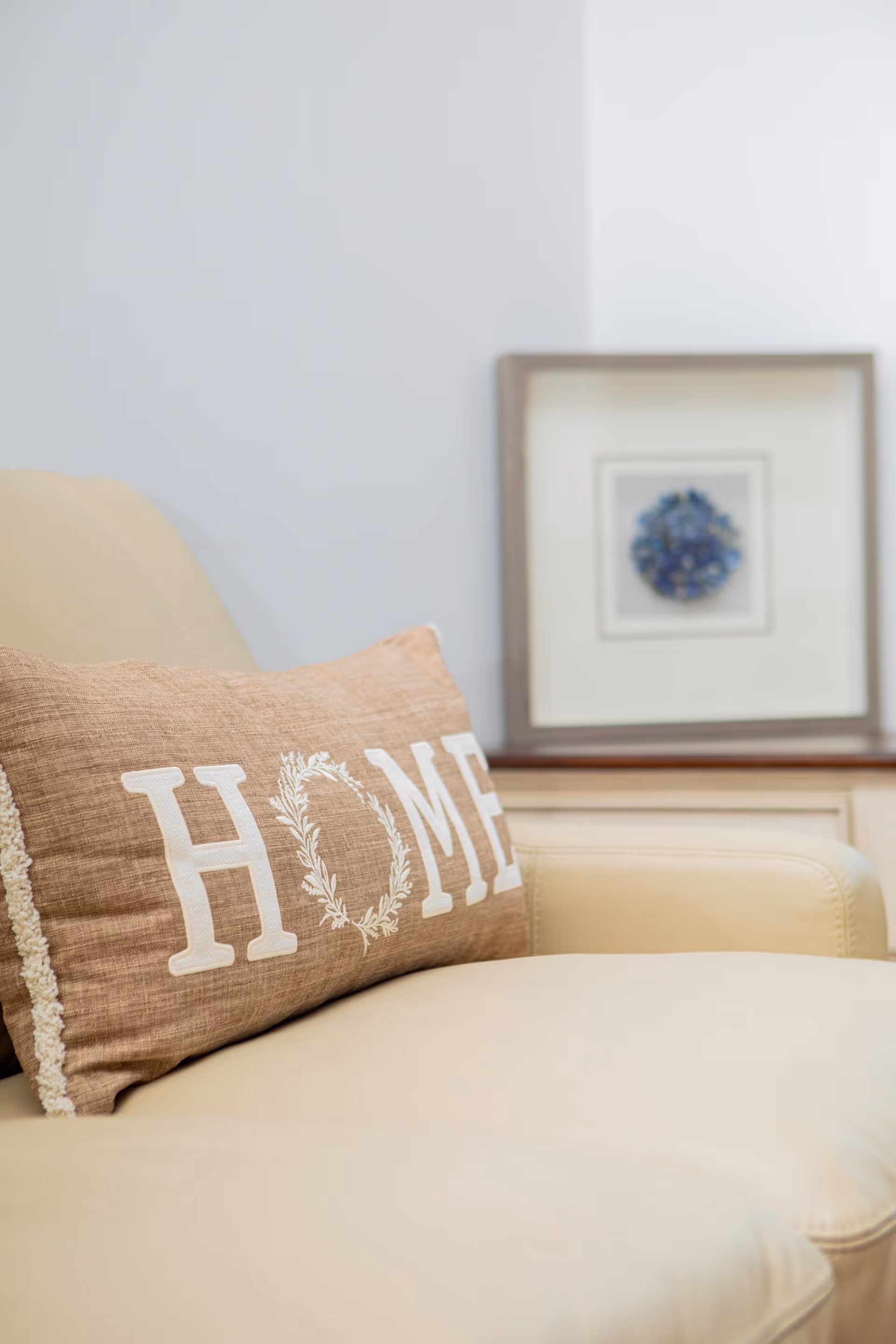 Close-up of a beige leather sofa with a brown decorative pillow that has the word 'HOME' embroidered on it. In the background, there is a framed piece of artwork resting on a piece of furniture against a white wall.