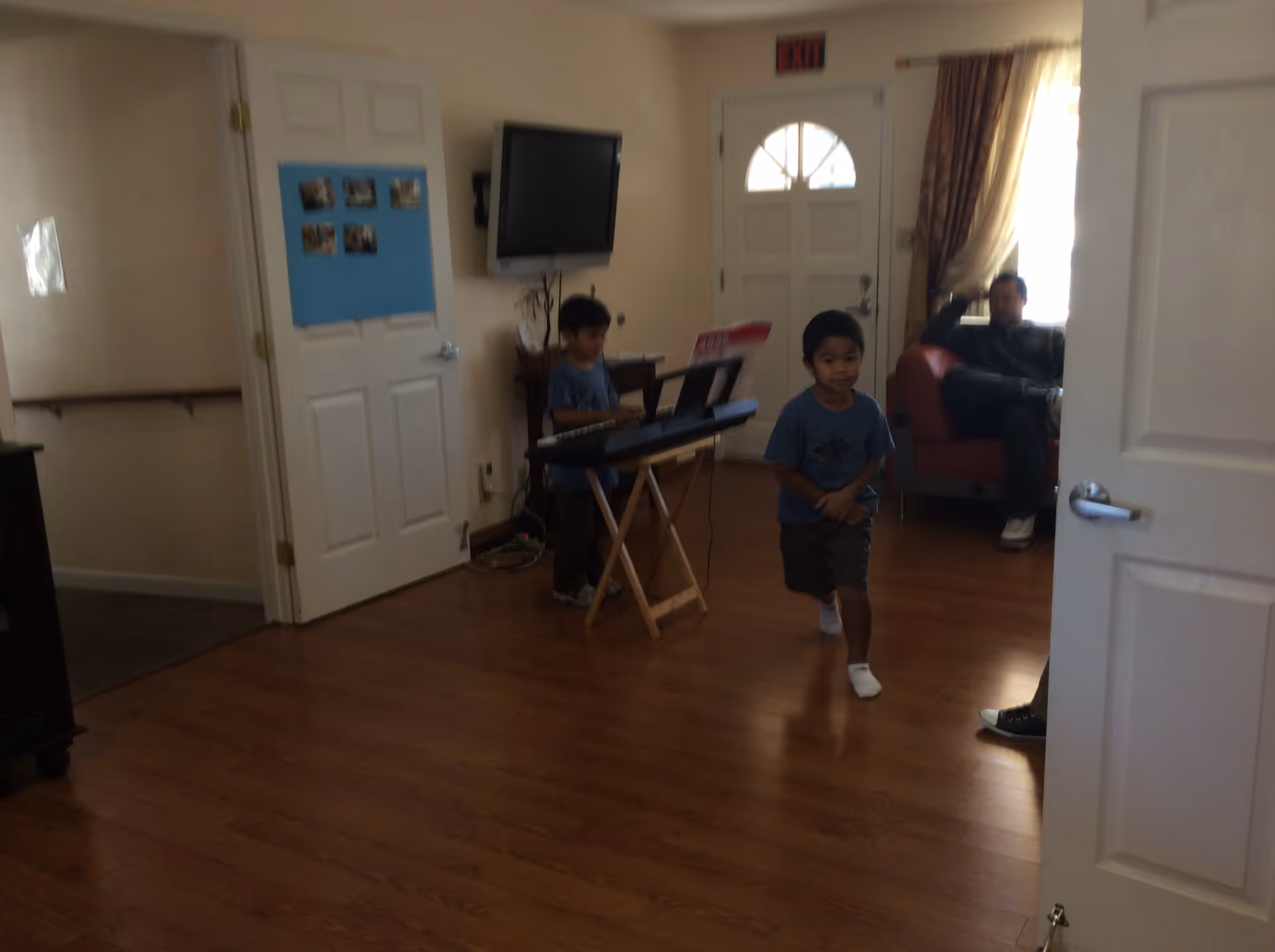 Interior room with wooden flooring, a white door with a blue bulletin board, a wall-mounted TV, a keyboard on a stand being played by a child, another child walking in the room, and an adult sitting on a red armchair near a window with curtains.
