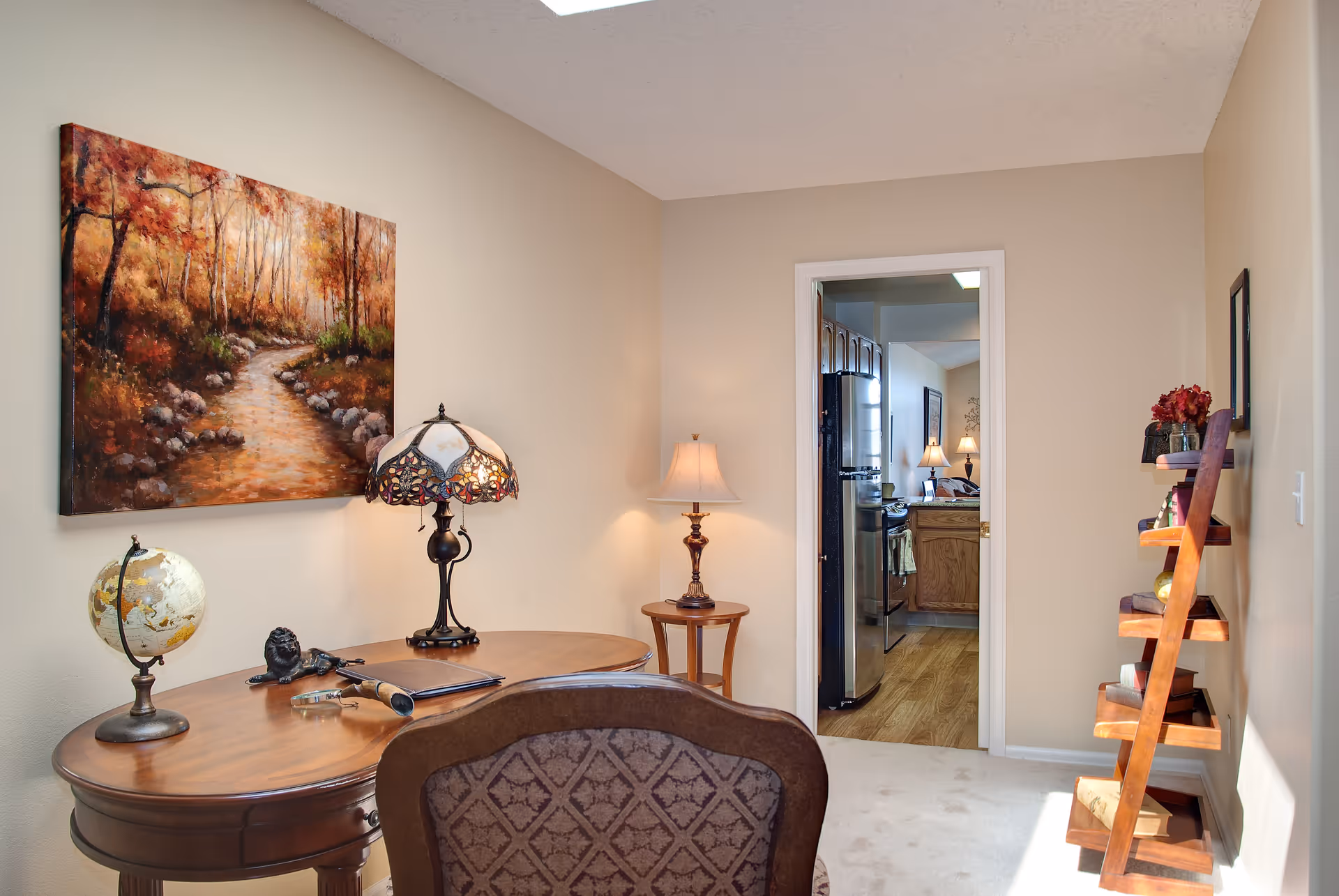 A cozy interior room featuring a wooden desk with a decorative lamp, a globe, and some small items on it. A patterned chair is positioned in front of the desk. On the wall above the desk is a large painting of a forest pathway with autumn foliage. To the right, there is a wooden ladder-style shelf holding various decorative items. A doorway leads to a kitchen area with wooden cabinets and stainless steel appliances. The room has beige walls and carpeted flooring.