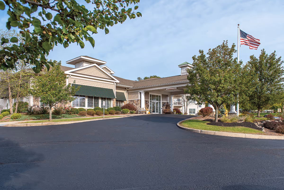 Exterior view of The Addison of Parklands senior living facility showing the main entrance with a covered driveway, landscaped greenery, trees, and an American flag on a flagpole against a partly cloudy sky.