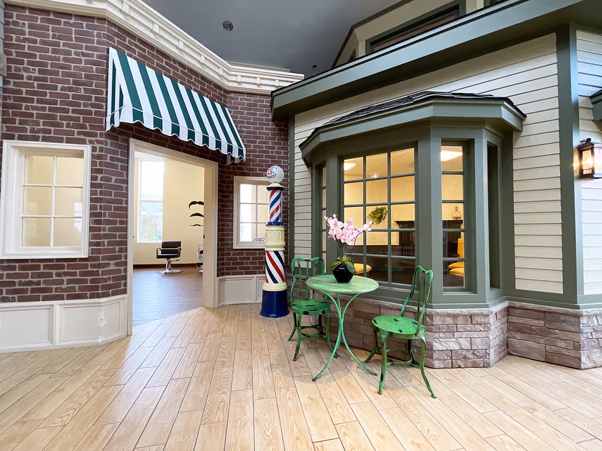 Indoor scene resembling a small village street with a brick wall and green-striped awning above a doorway leading to a room with salon chairs. A traditional barber pole stands near the entrance. To the right, a bay window with green trim looks into a cozy room with a fireplace and yellow chairs. In the foreground, a small green metal table with two matching chairs and a vase with pink flowers sits on a wood-patterned floor.